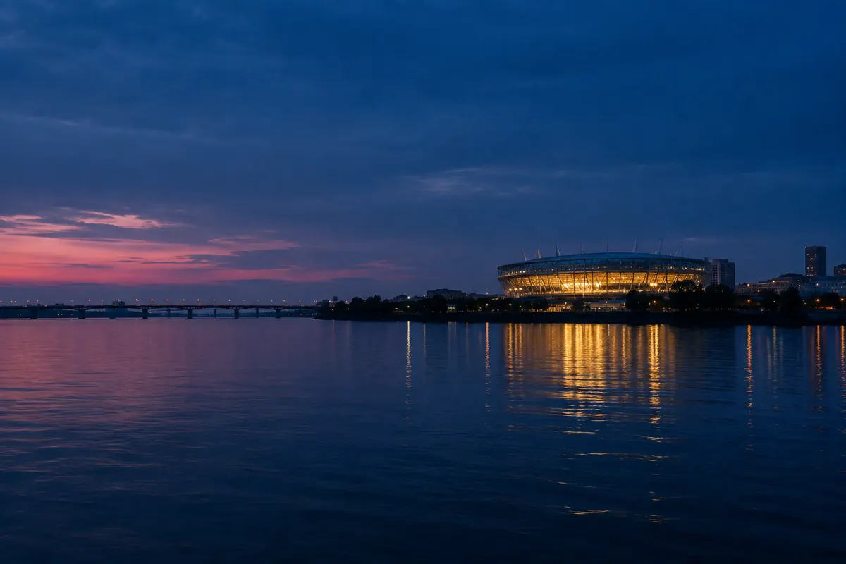 A single lit sports arena on the far bank of a wide dark river at blue hour, its lights reflected in the water beside a distant bridge