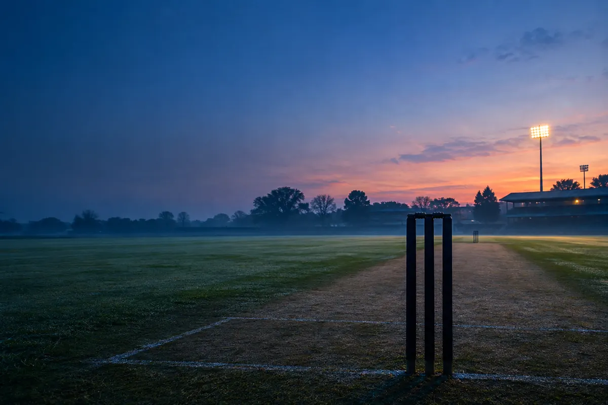 A cricket pitch at dawn seen from behind the stumps, the worn strip stretching away across dewy grass under a thin rose glow