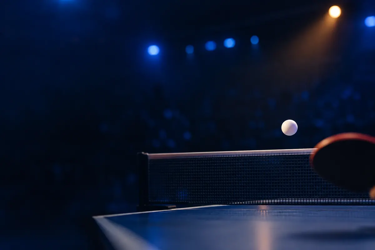 A table tennis ball frozen in mid-air just above the net, the blurred edge of a paddle in the foreground, a dark arena beyond