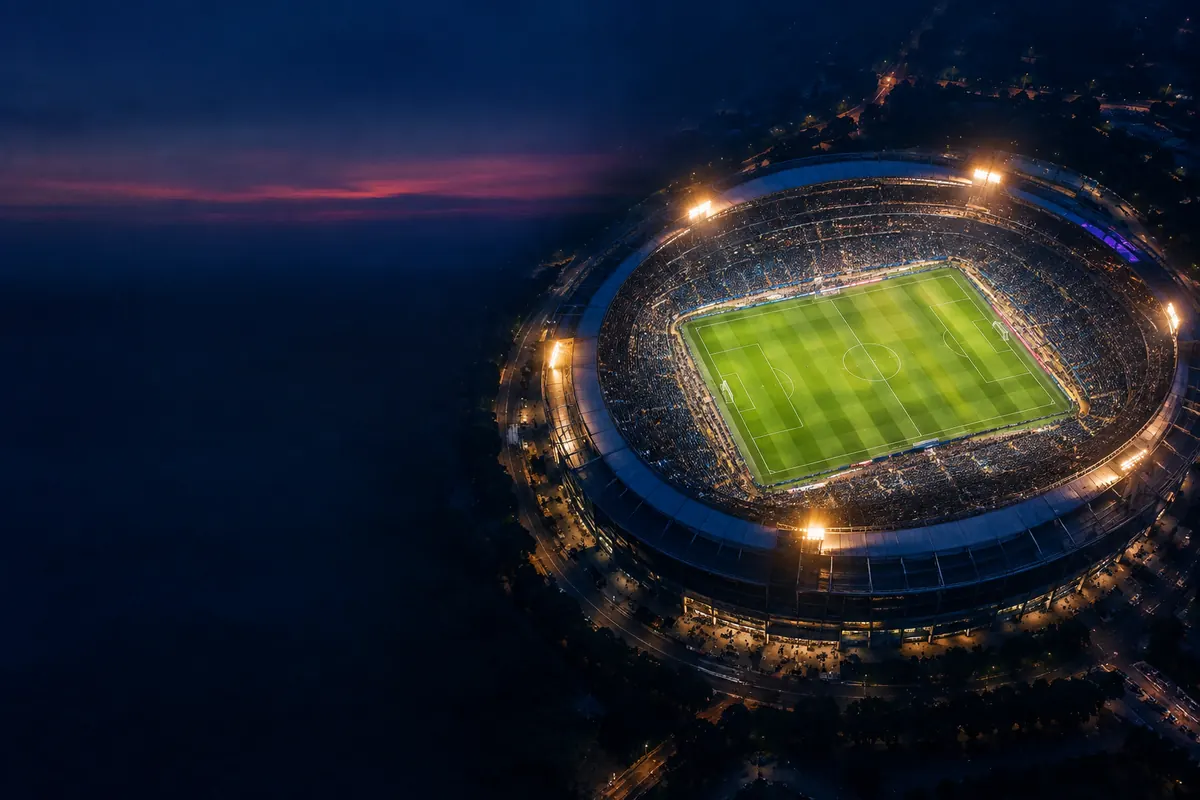 Aerial night view of a packed oval stadium, tiny points of light around a glowing green pitch, warm amber floodlight flares