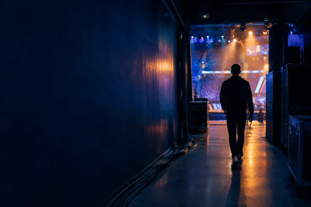 Dim backstage corridor of an esports arena, a silhouette walking toward a lit stage.