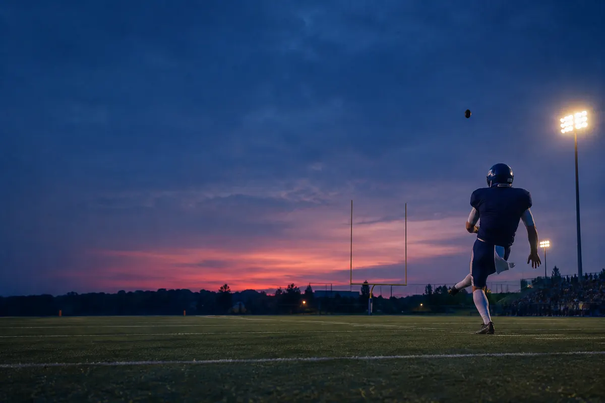 An American football kicker seen from behind following through after a field goal attempt, the ball rising toward the posts at dusk