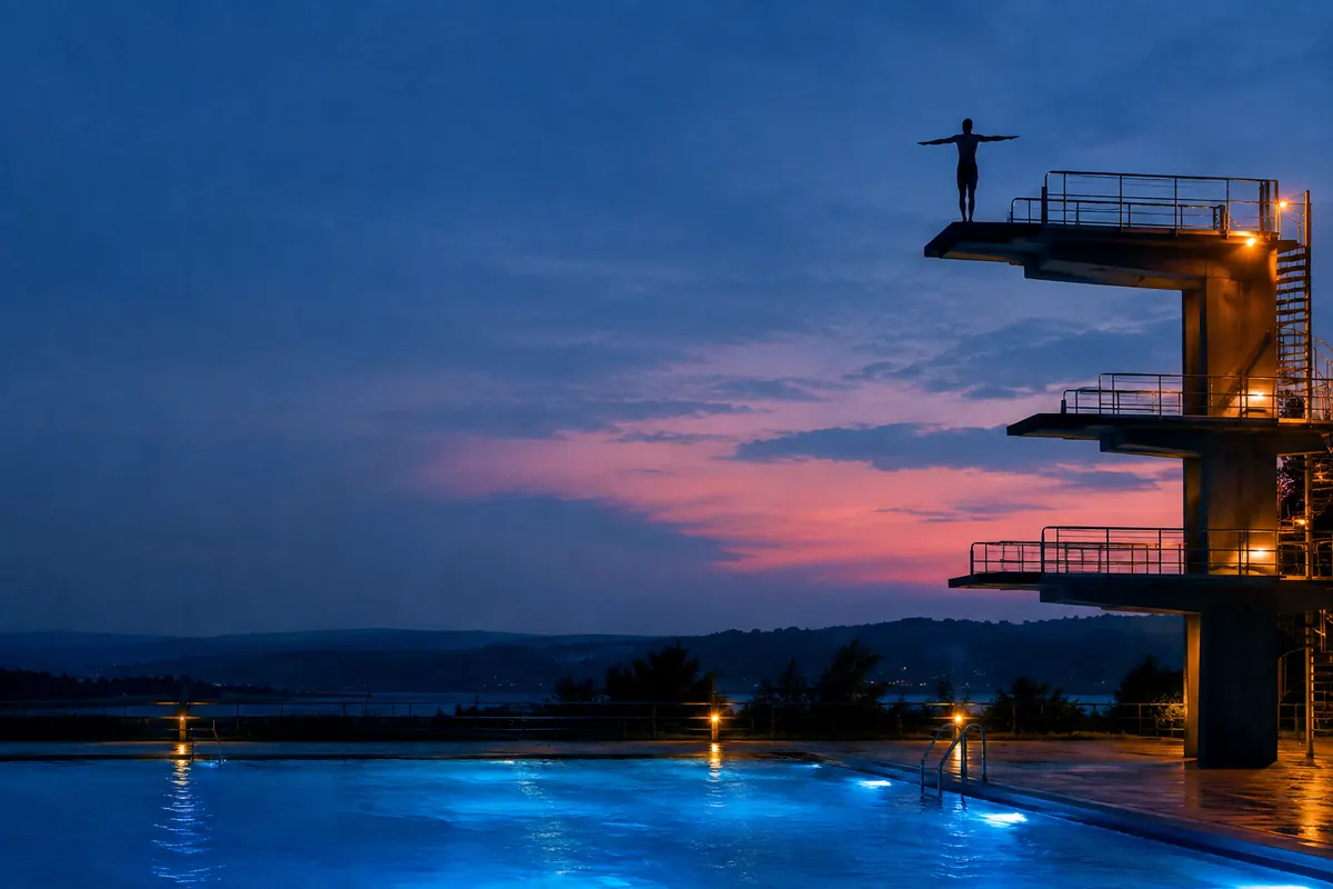 A high diver in silhouette at the top of a diving tower at blue hour, arms out, the pool glowing cobalt far below.